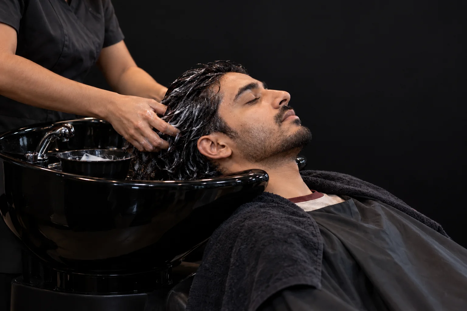 A client resting at a black shampoo basin while conditioner is worked through his hair against a plain charcoal background
