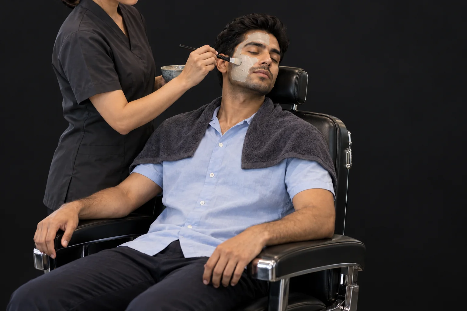 A client resting in a salon chair during a facial mask application against a plain charcoal background