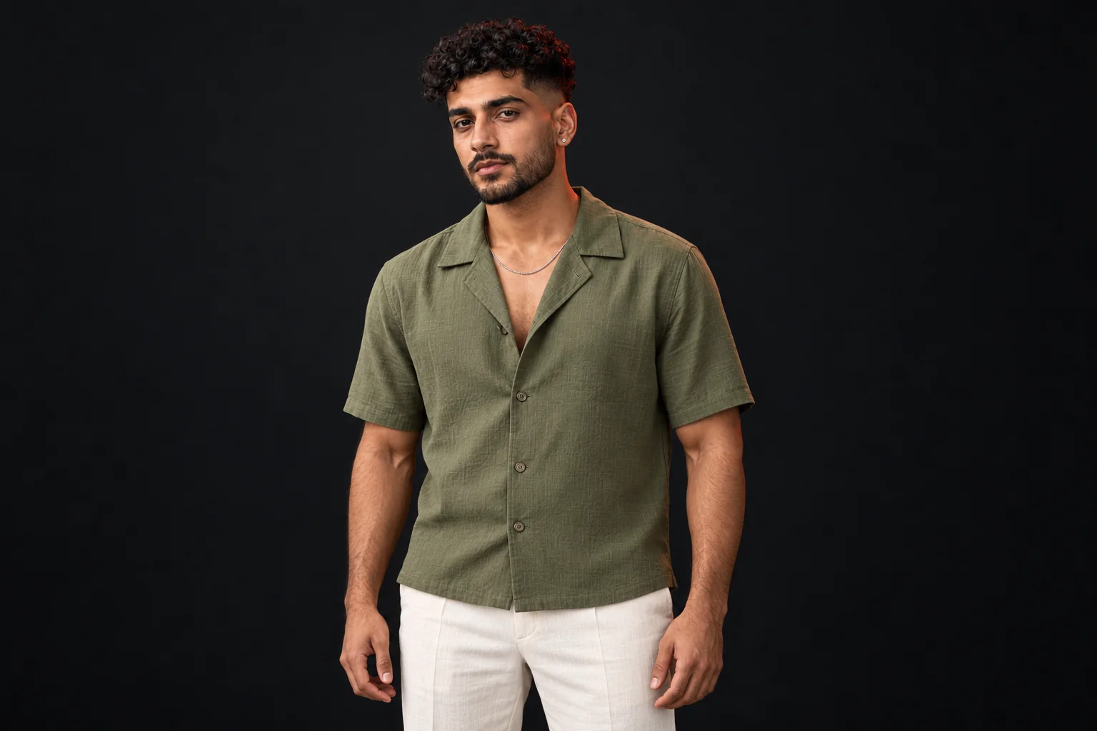 Man standing upright with a curly crop, low fade and short beard, wearing an olive shirt and cream trousers